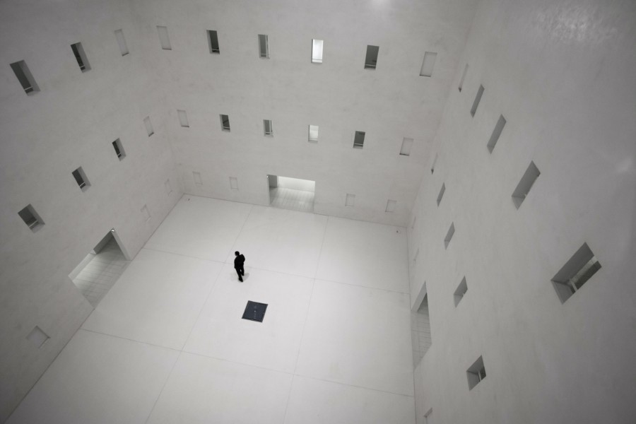 An entirely white room viewed from high above where a silhouette of a person stands in the near centre and looks at all the door shaped openings around the room at different heights, choosing which one to go through.