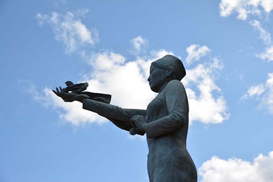 A photo of a large metal statue, viewed from below with a blue sky behind it. The statue is of a woman with an outstretched arm that a bird has landed on.