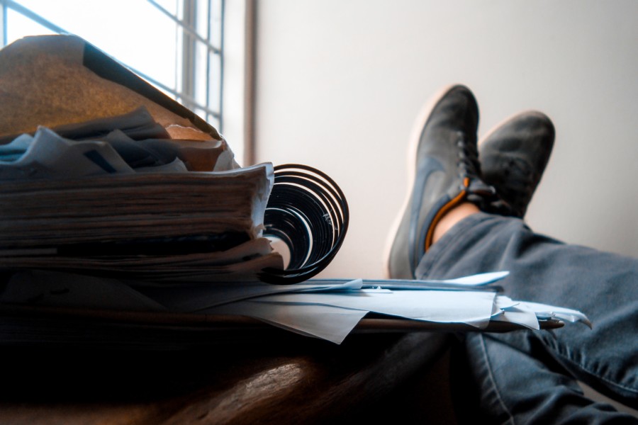A person rests their feet on a desk next to a pile of papers and notebooks.