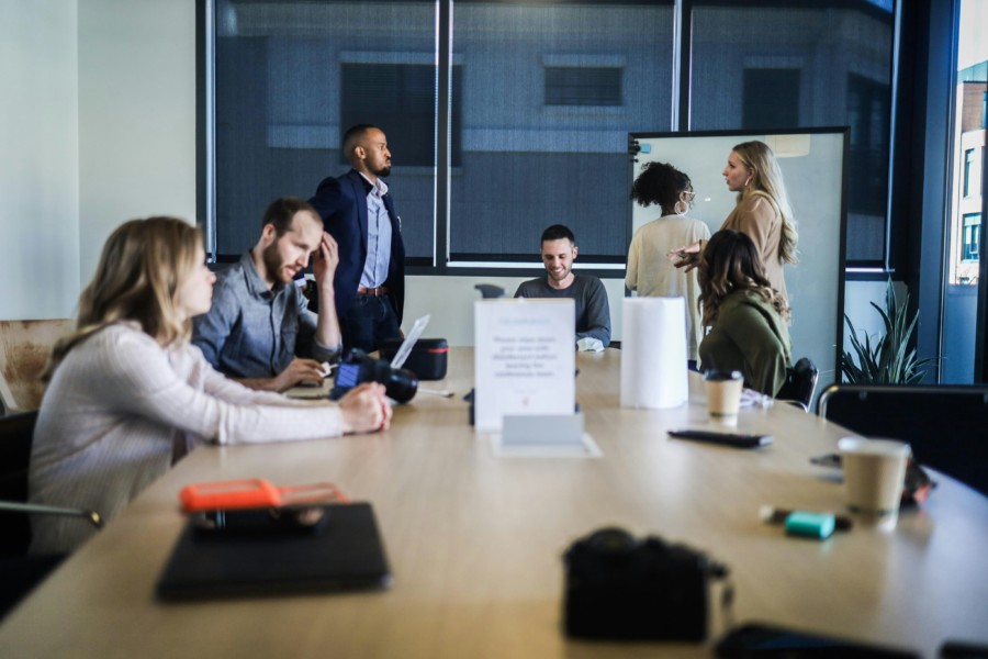 A photo of people in a office meeting room, some stood and some sat. It appears to be the end of a meeting where more informal discussion and closing is occurring.