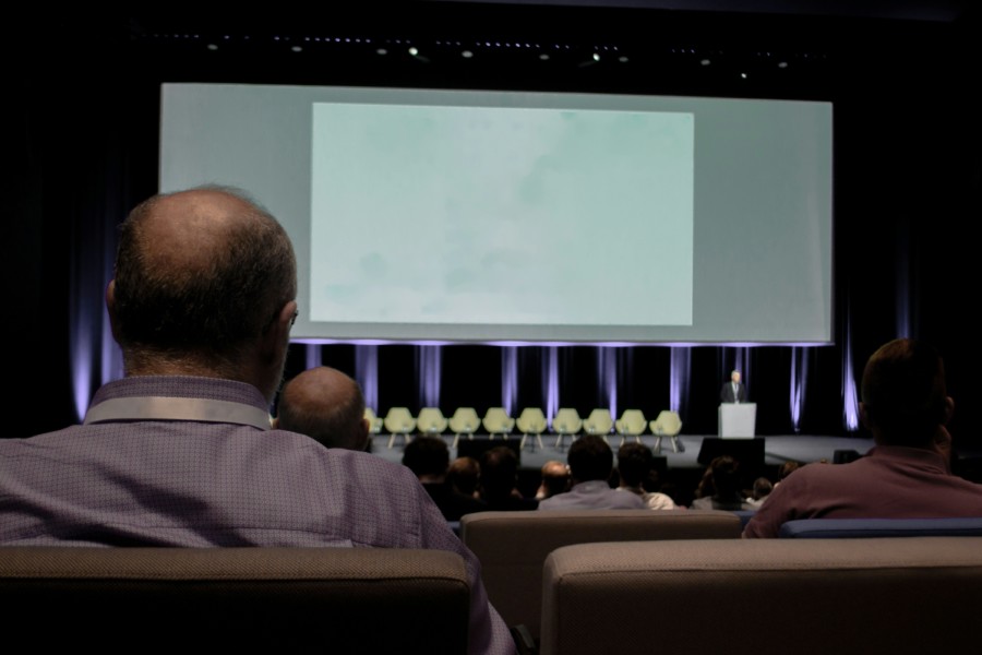 A person viewed from behind sits in the lecture theatre seating looking up at a presentation screen and the stage filled with chairs and a solo speaker below.