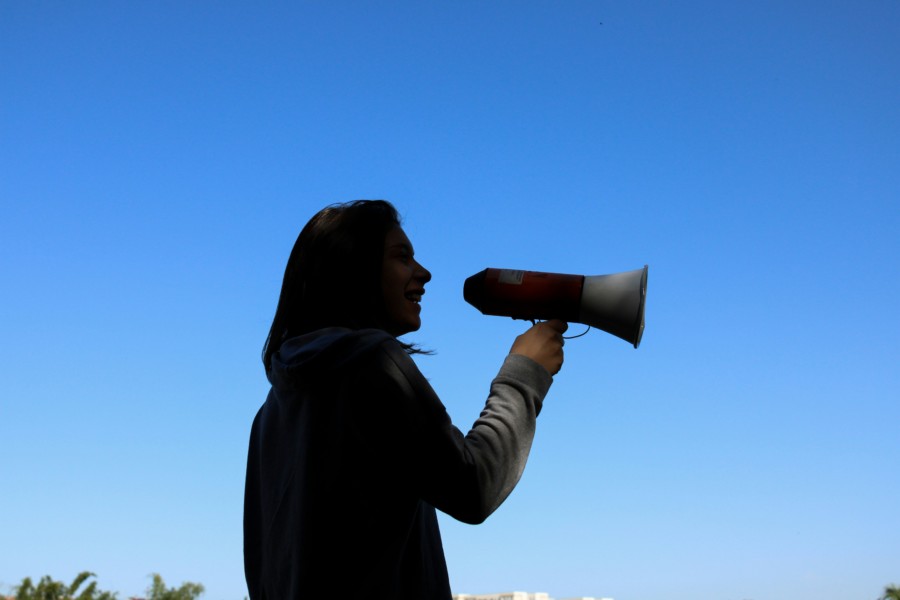 A person in silhouette against a bright blue sky holds a megaphone to the right and speaks through it.
