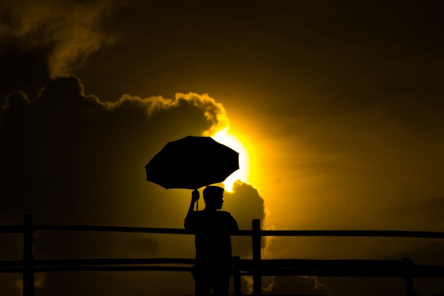 A photo of a silhouetted person holding an umbrella against a yellow sun peeking out from some dark and stormy clouds.