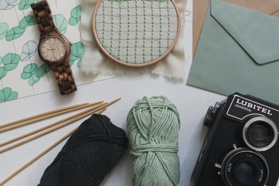 A photo of a collection of pale green and black items viewed from above. There is a camera, some knitting wool, an embroidery hoop with tiny gingham fabric, a brown watch, a pair of envelopes and some patterned paper underneath.