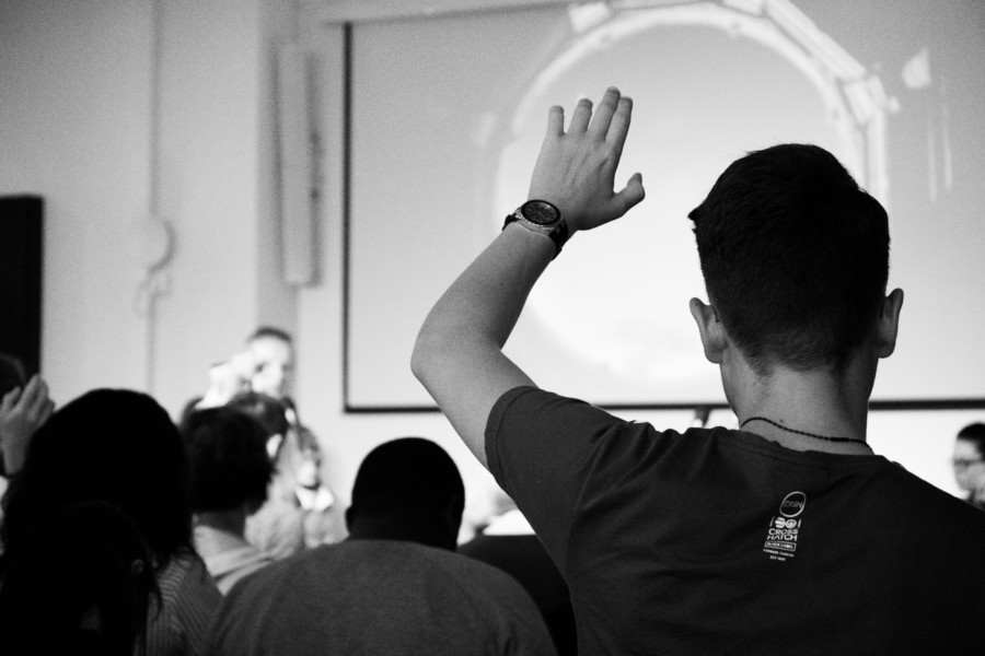 A black and white photo of the back of a guy raising his hand to ask a question in a lecture or presentation.
