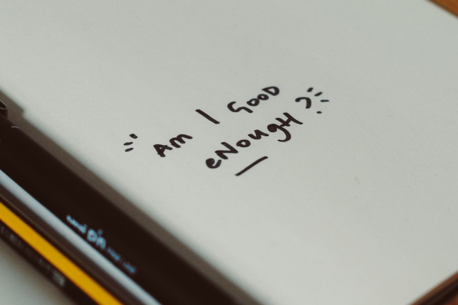 A photo of a white notebook open on a page with a pen and a pencil resting in the open spine. The page is largely empty except for a small question written in black that says: "AM I GOOD ENOUGH?"