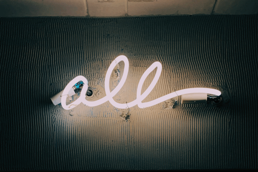 A photo of a dark room with a neon sign that reads "all" in lowercase handwritten script.