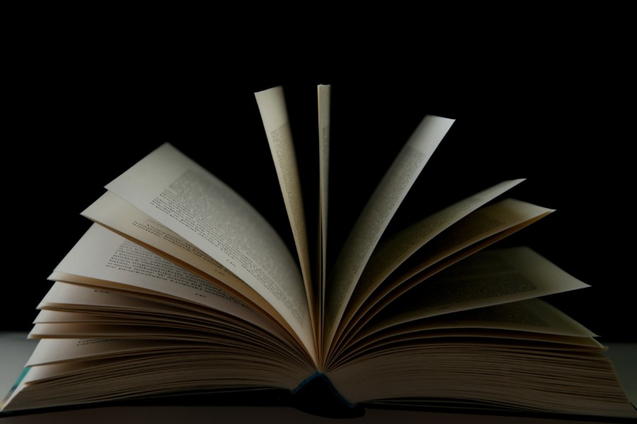 A book is open in front of a dark black background where the pale fanned open pages hover in the air as someone has flicked through