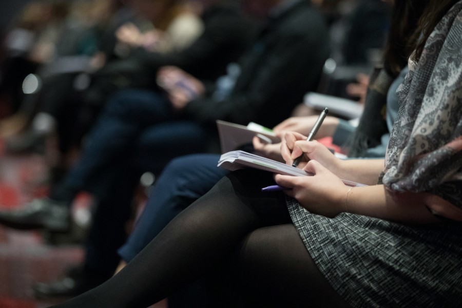 A close-up photo of a person seated in a row of people writing in a paper notebook on their crossed knees. The rest of the row of people are blurred in the background with high focus in the foreground. The scene suggests taking notes in a conference or lecture with smart casual attire. No faces are visible.