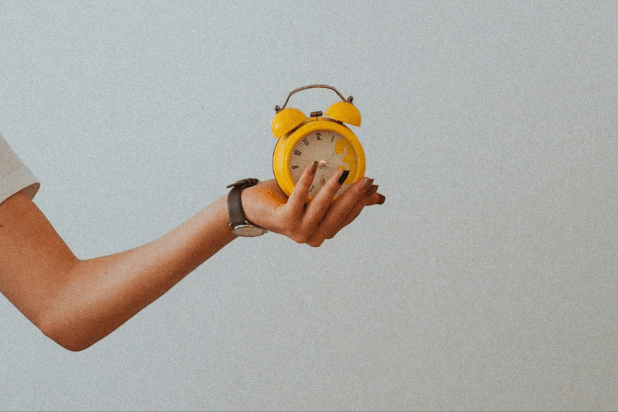 A person in a white t-shirt holds a bright yellow traditional alarm clock facing towards the camera.
