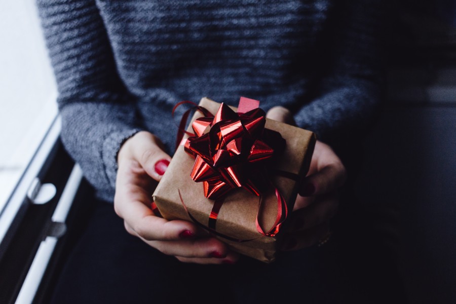 A person with red nails and grey jumper holding a brown paper wrapped present with shiny red rippon tied in the traditional cross and bow way with a sticker ribbon rose stuck on top. Subtle meaning of rediscovering the joy in your research over Christmas break as you do when receiving and gifting presents.