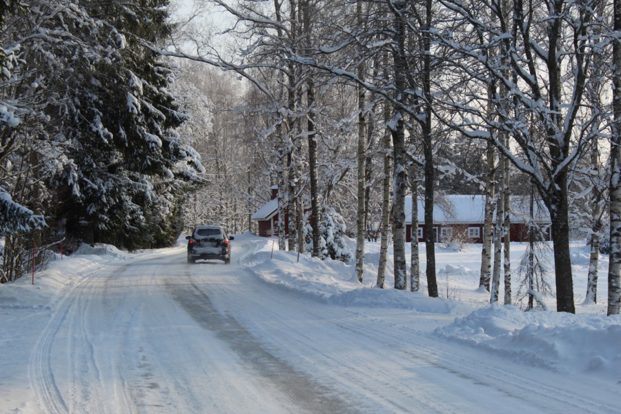 A car drives slowly on a heavy snow and ice filled road between lines of tall forest trees.
