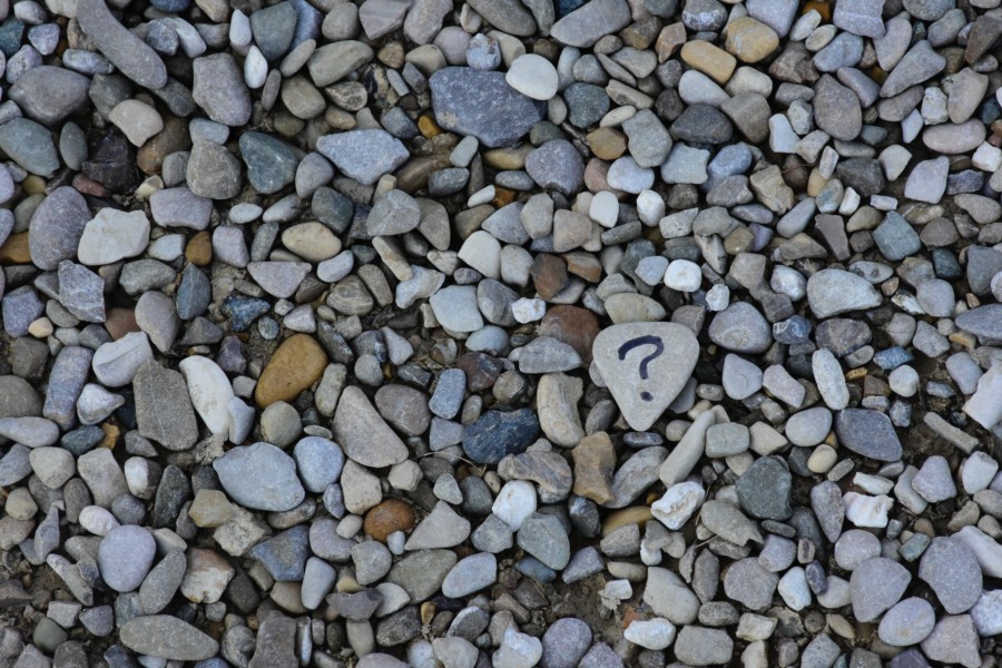 A photo of a pebble beach with a variety of greys, white, orange and blue grey pebbles in small sizes. A slightly larger one in the righthand third of the image has a question mark drawn on its face in black marker pen