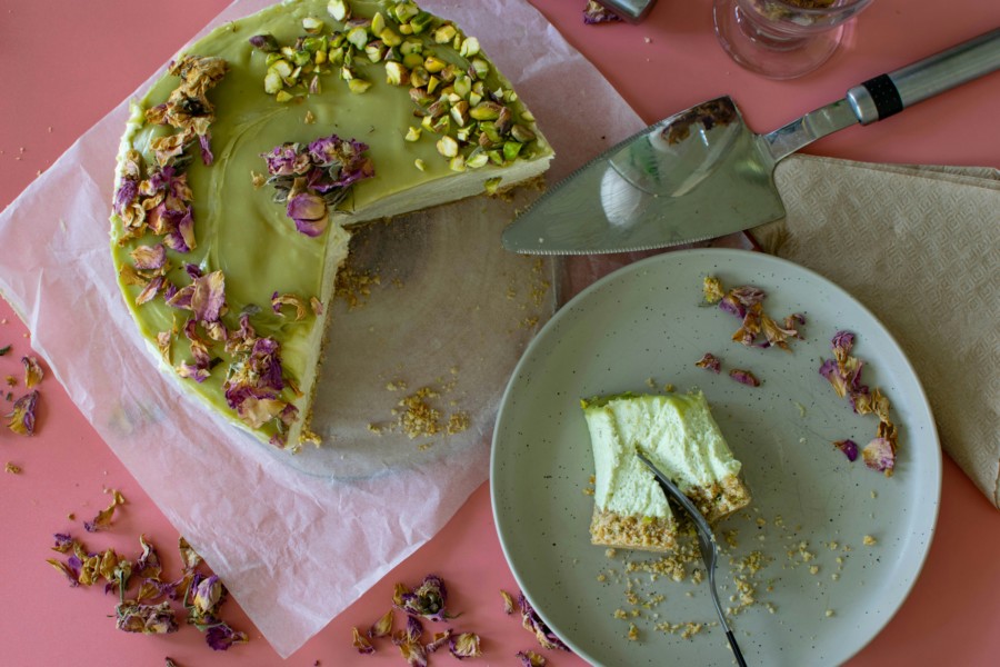 An overhead photo of a pale green pistachio cheesecake with a large segment cut from it and half eaten on a nearby plate. The cake is decorated with pistachio crumbs and petals, while sitting on a bright pastel pink surface.