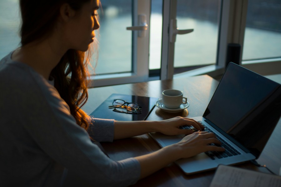 A woman sits at a desk near a window with a laptop and a coffee mug.