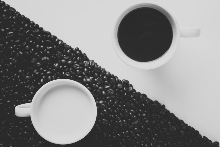 2 white mugs sit in opposite corners of an image where the one to the bottom left is surrounded by perfectly arranged dark coffee beans and holds a white liquid, while the other mug holds a dark liquid and is only sat on a pure white surface showing the contrasts of dark and light