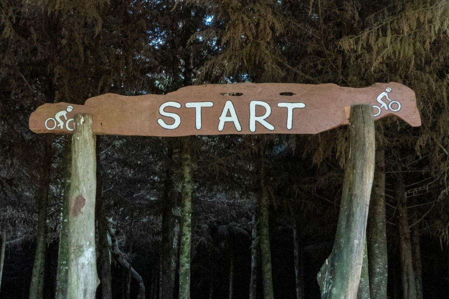 A wooden sign made from a flat slice of tree trunk held up by a big branch each side and stuck int the ground of a forest setting. On the flat part, the word "START" is painted in white which signals the beginning of a cycling route, with little cycling illustrations either side of the word.