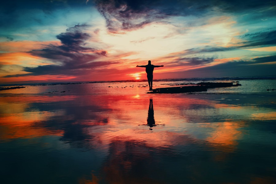 A silhouette figure with outstretched arms stands on a small sandbank of a beach where the surrounding sandy water reflects the vibrant colours of the sunset to fill the whole image. There's purple wispy clouds, pinks, oranges, blues and reds all mixed in to the dusky sky.