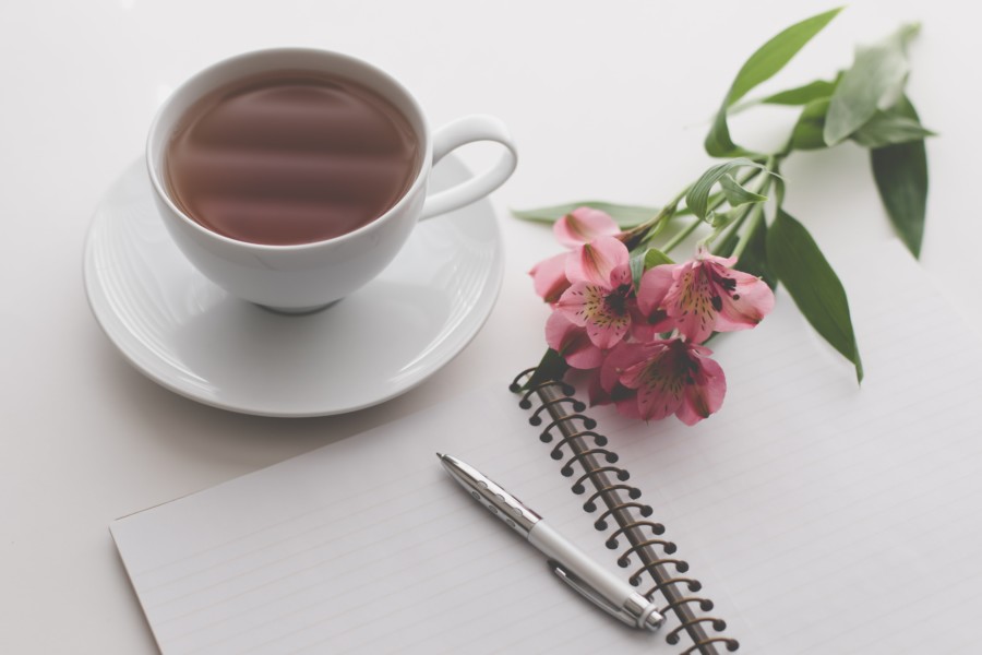 A white cup full of tea and saucer sits next to an open ring-bound notebook with a silver pen on top. Lay across the notebook is some pink flower cuttings with stems and leaves.