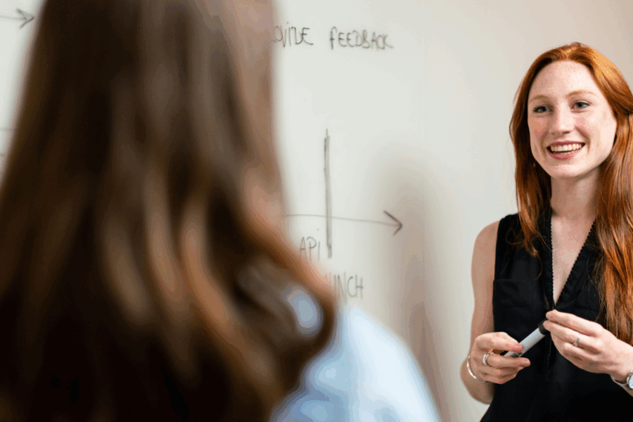 2 people speak in front of a whiteboard. One faces away from the camera towards the other who is holding a marker and we can assume she is a lecturer or teacher. The teacher has long red hair and wears a formal black waistcoat and smart black trousers.