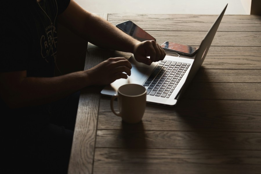 A photo of a person sitting at a wooden table with hands hovering over a laptop keyboard in a dark shaded room. A mug sits nearby casting further shadows.