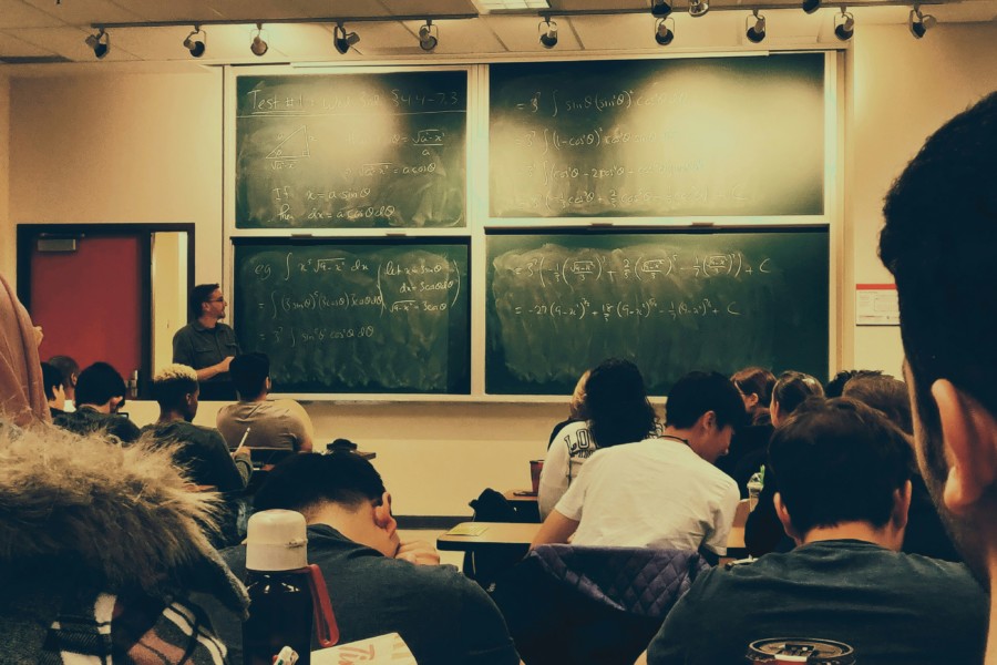 A photo of a classroom from the perspective of a student looking up at a classic green chalkboard of notes and a teacher stood nearby. The image has had a yellow tint filter added.