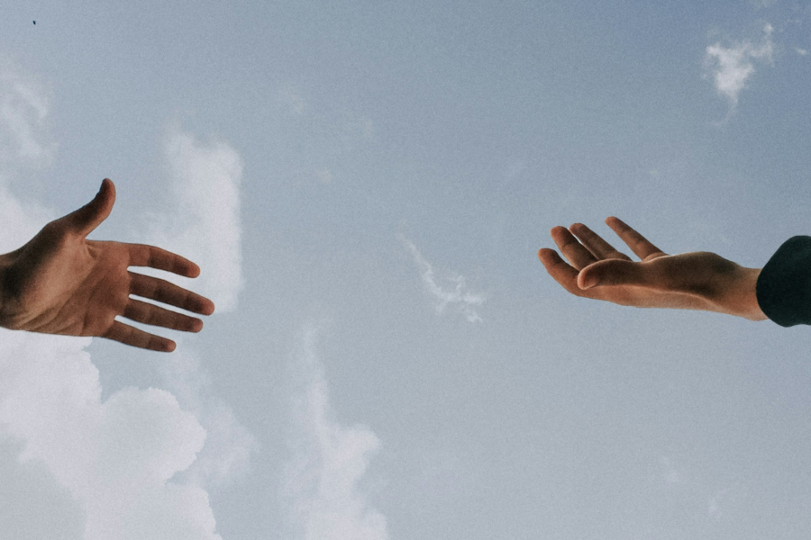 A photo of two people reaching out their hands to each other but not quite touching with a pale blue cloudy sky in the background