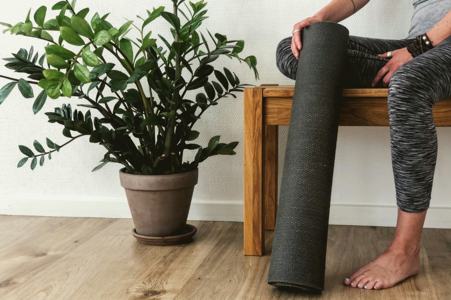 A person sits on a bench in a yoga studio holding their rolled up mat upright. There's a large potted plant nearby in the bright airy space.