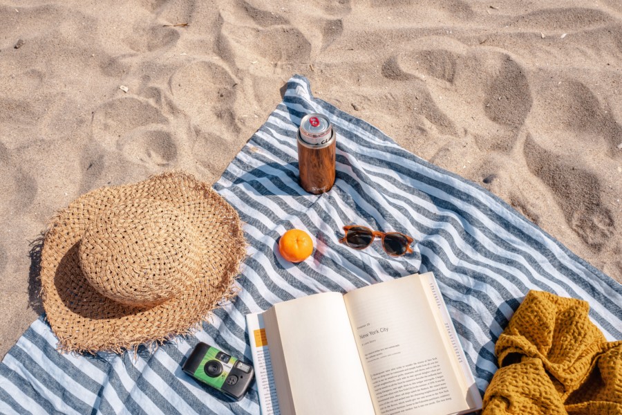 A blue and white striped picnic blanket sits on a white sandy beach with a straw hat, an open book, a disposable film camera, a pair of sunglasses, a mustard yellow cardigan, a small satsuma and a canned beverage in a cooler displayed on top.