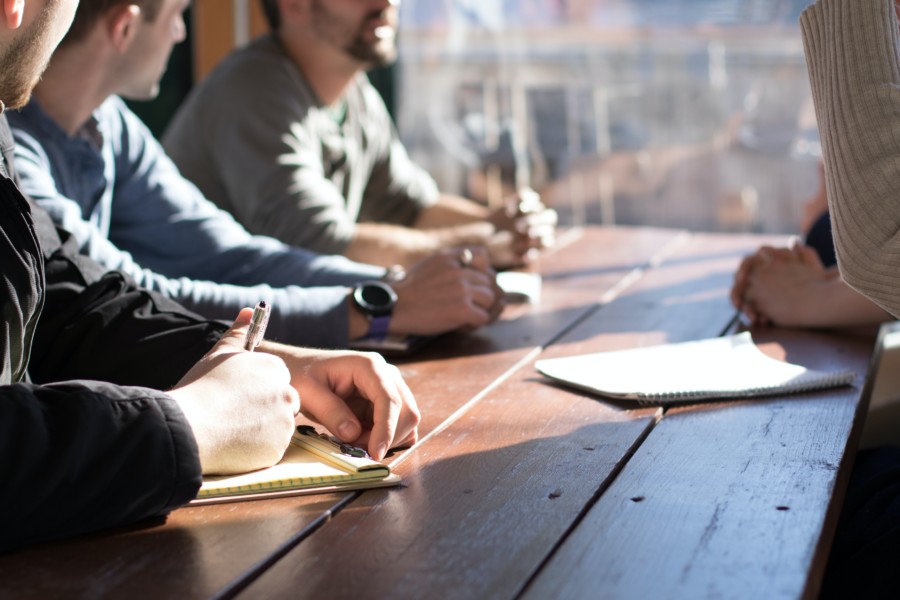 People in a meeting gathered around a wooden bench table with notepads and faces cropped out of the image. the sun shines on their hands and the table illuminating the discussion.