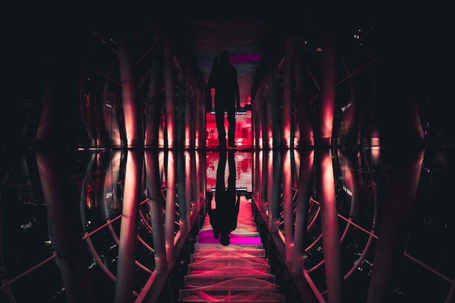 A dark silhouette figure walks down a central glass walkway that reflects the large architectural ceiling joists to create an ominous scene. The colouring is dark pink, red and orange evoking images of dark, shady behaviour.