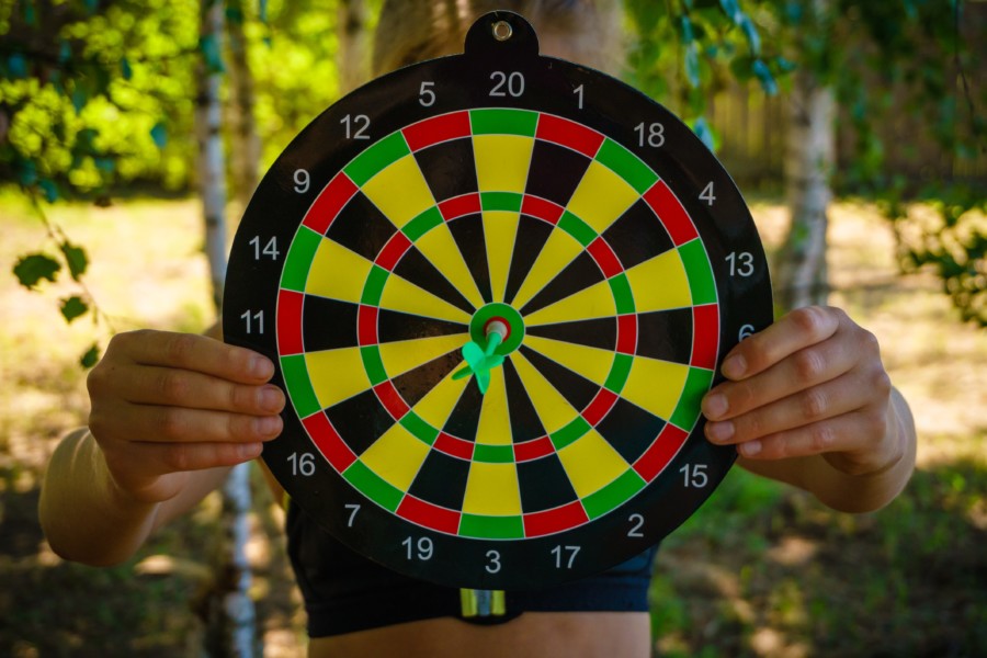 A person holds up a colourful circular dart board to the camera where a green dart is perfectly positioned on the bullseye in the centre.