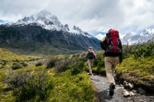 2 people in hiking gear hike up a rocky path towards a snow capped mountain surrounded by grassy plains.
