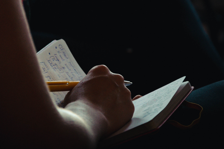 Person writing notes in a notebook with a pen, in a dimly lit space, probably listening to a speaker out of view.
