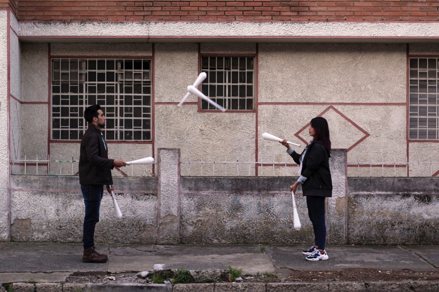 2 people juggle a set of white batons between each other a few paces apart in an urban area.