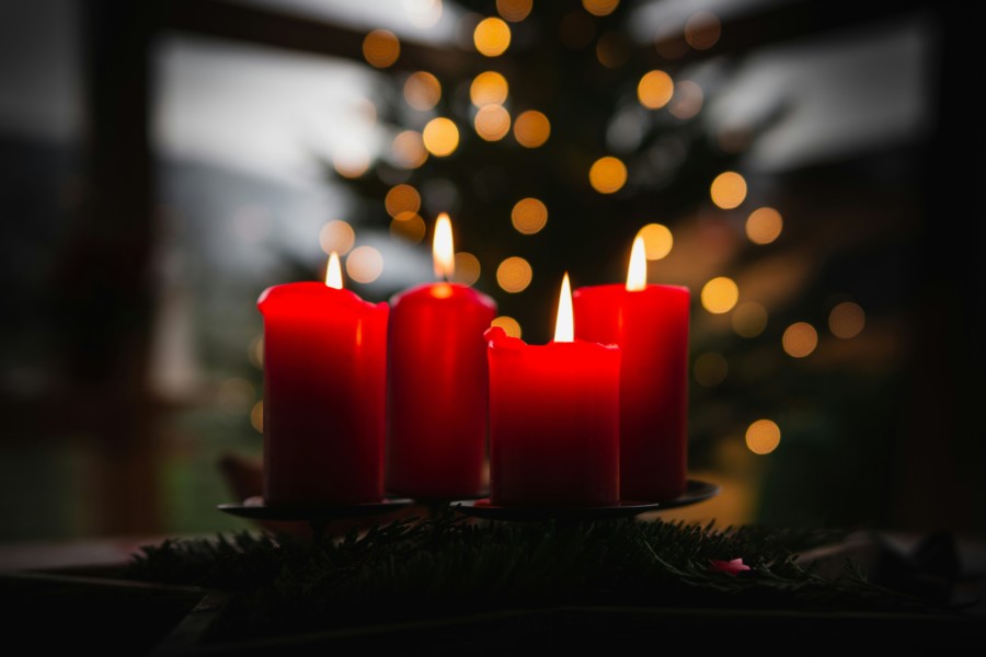 A group of bright red pillar candles are lit in the foreground with an unfocused background of fairy lights adorning a christmas tree.