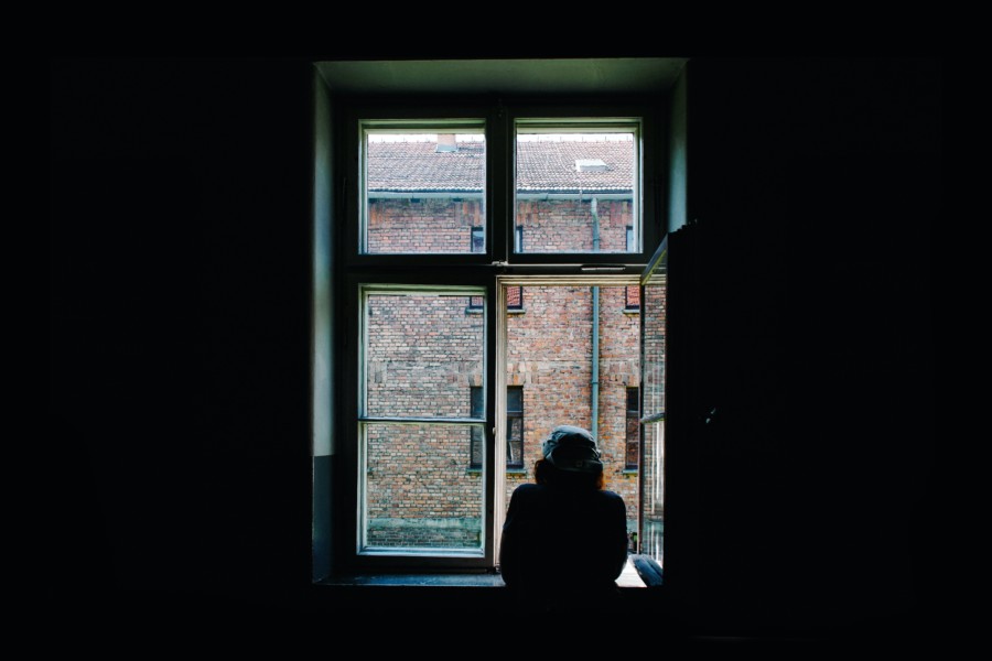 A person in a dark room stands at and looks out of an open window in a hunched position giving a sense of sadness or in deep thought.