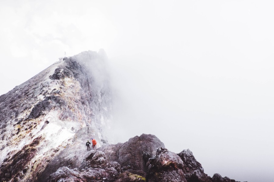 A photo of mist wrapping round a mountain ridge. There are two hikers on the middle ridge where a false summit is before them, followed by a higher peak in front of them that stretches up into the cloud.