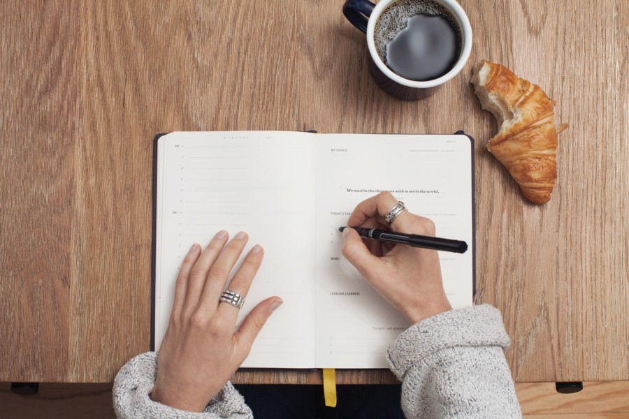A person in grey knit writes in a notebook on a wooden table with a black coffee mug and a croissant with a bite taken out of it to the side.