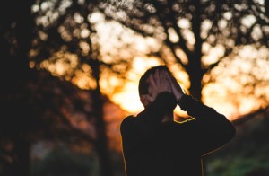 Person puts both hands over their face in a distressed gesture against a bright orange sunset coming through some dark trees in the background