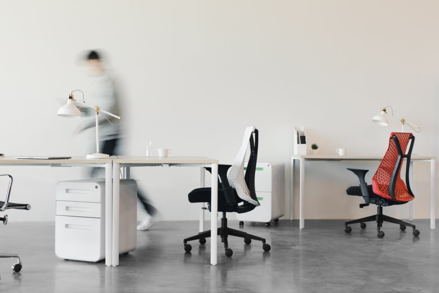 A stark white office scene with 2 white desks and an ergonomic desk chair pulled underneath each. They are both empty but a person walks quickly across the room creating a blurred rushed effect via the camera's slow shutter speed.