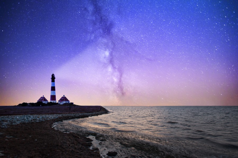 A dusk scene where a lighthouse on the rocks beams a ray of light out to the seascape beyond where the sky is full of purple, blue, pink colours and a galaxy is visible.