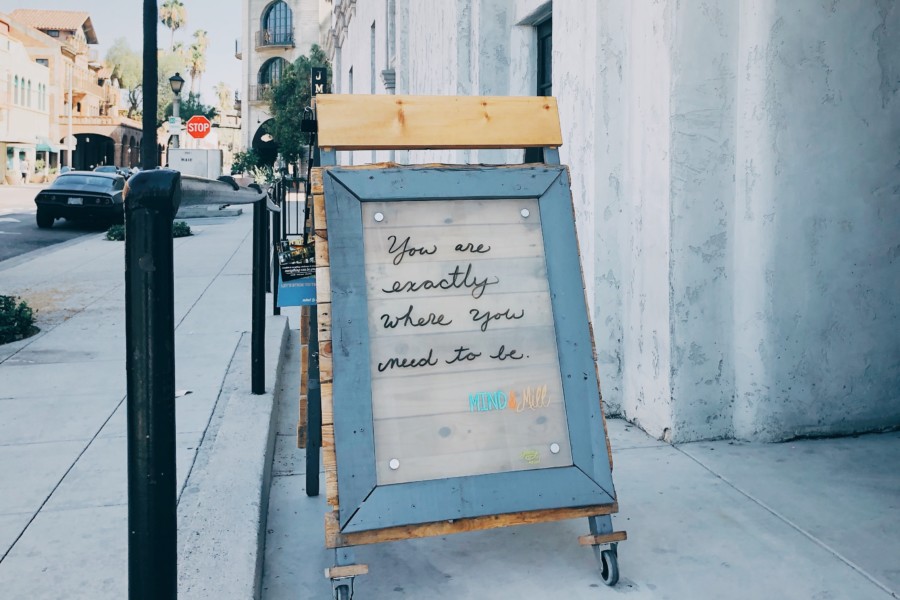 Sandwich board style wooden sign on the pavement outside a coffee shop that has a handwritten quote on the paper inside the glass waterproof screen with "you are exactly where you're supposed to be"