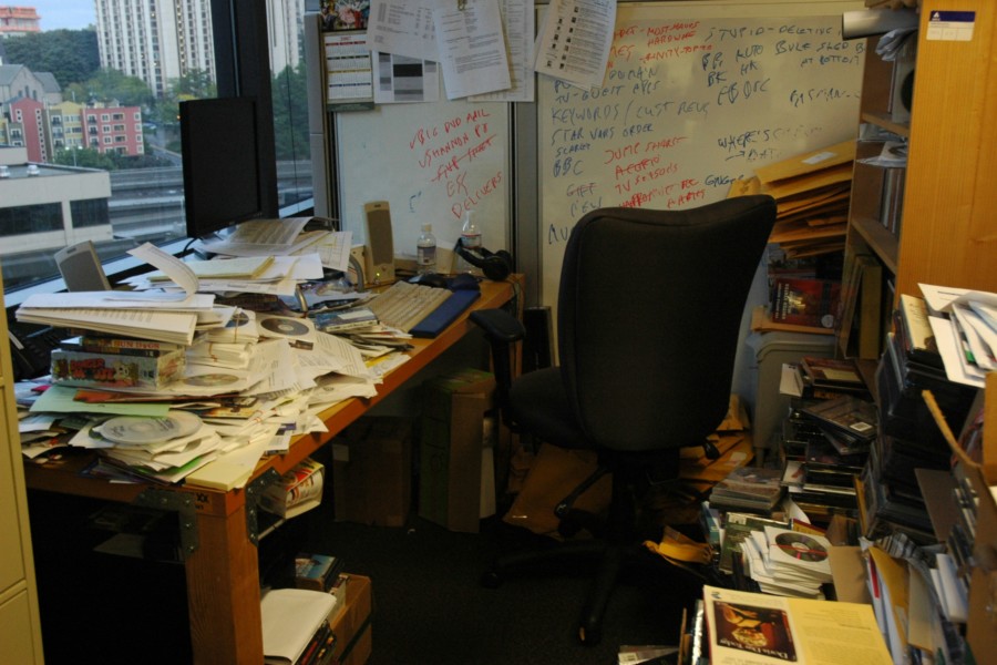 A photo of an academic office where every surface of desk and bookshelves is piled high with various books and papers, CD's and clutter.