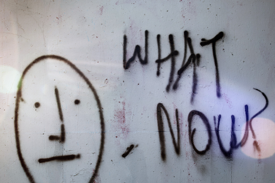 Some black graffiti on a white brick wall that says "What Now?" in capital letters next to a simplistic neutral face that has a horizontal line for a mouth