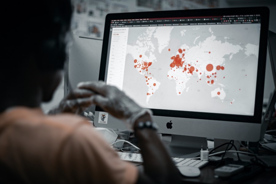 A person looking at a computer screen that is displaying a world map that shows red circles in varying sizes on the cities with large numbers of covid cases.