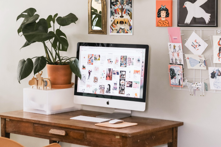 A home office set up with a collection of cute small posters and photos arranged in a loose grid of different sizes like a gallery wall with an iMac and pot plant on the wooden desk.