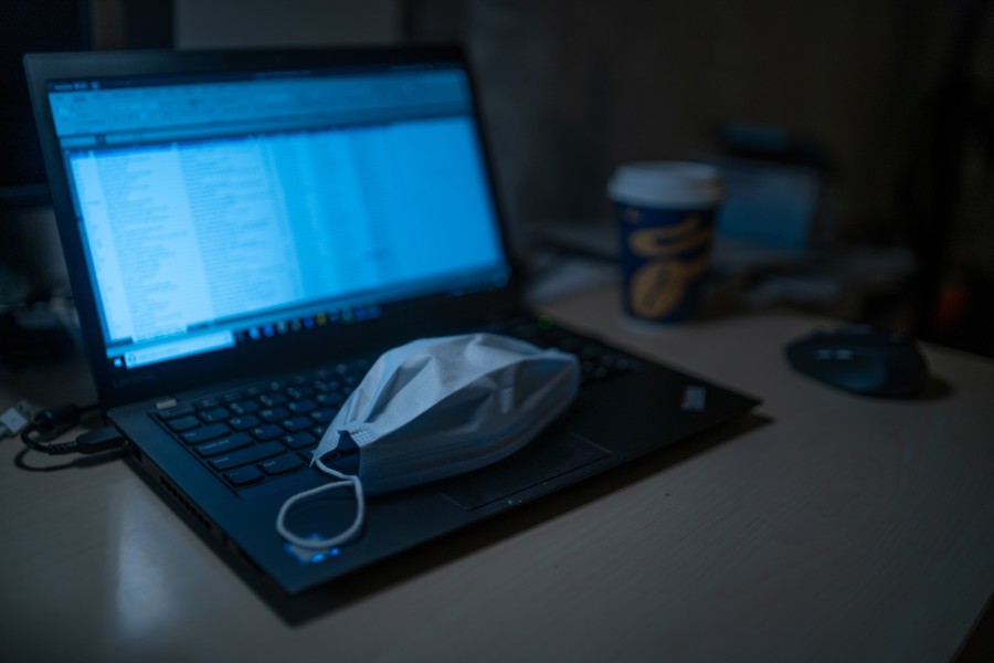 A laptop is open on a spreadsheet of data in a long vertical table where the blue light from the screen illuminates the dark room. There is a basic surgical mask left on the laptop keyboard and a takeaway coffee cup nearby on the desk.