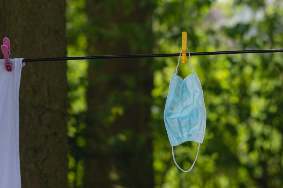 A blue face mask hangs from an outdoor washing line with green sunlit trees in the background