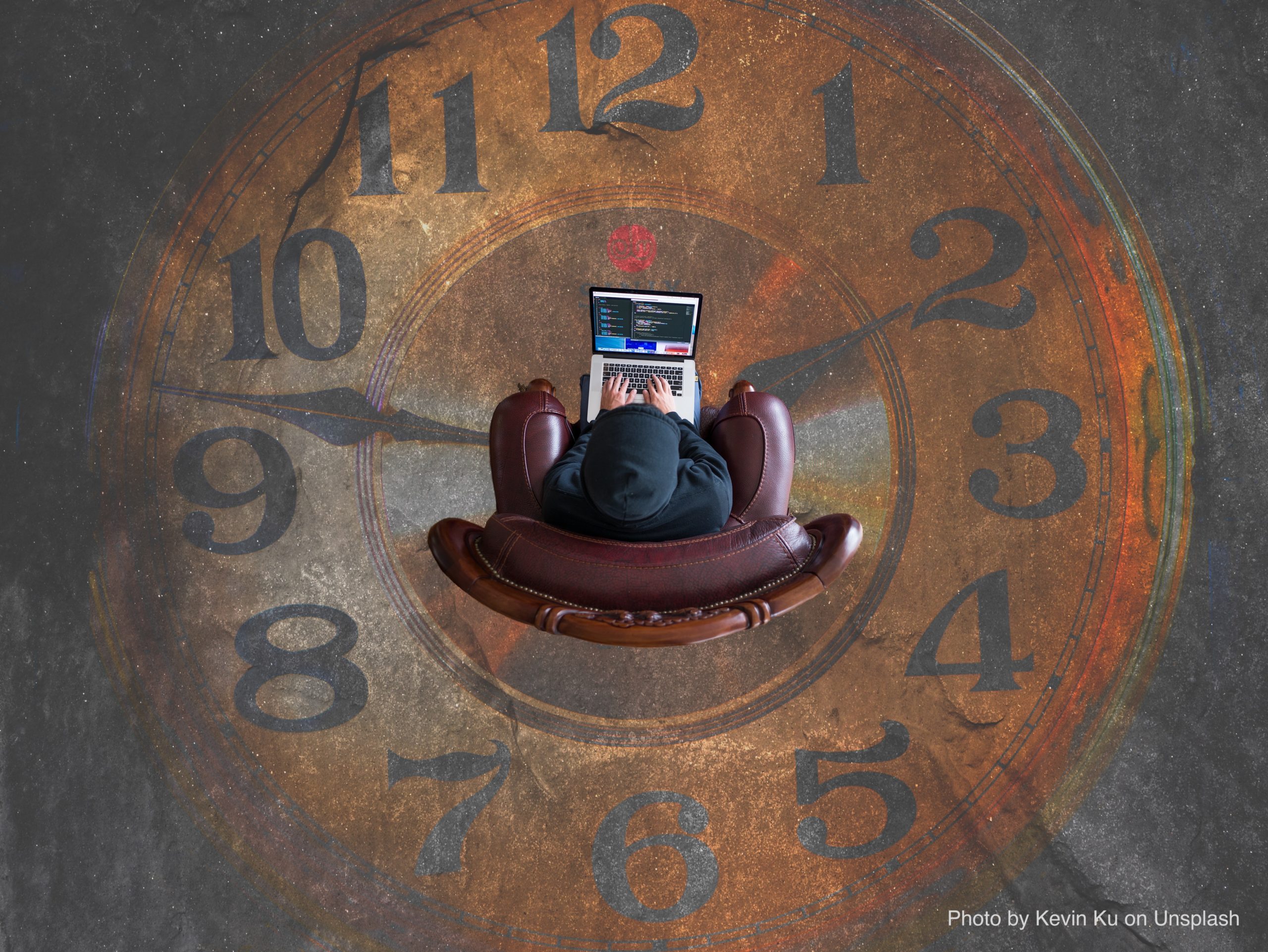 A person sits on a brown leather armchair working on a laptop which is perched on their knees. They are viewed from above and shown to be sitting in the middle of a rustic clock design that is displayed on the floor. The clock hands point to 10 past 9.